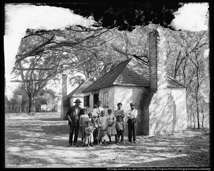 A family at the Hermitage, Savannah, GA. They stand in front of a small brick house with chimneys at both ends. The scene is set outdoors with large, leafy trees providing shade. The image is in black and white, and the setting appears to be historical.