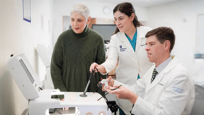Three medical professionals in lab coats demonstrate a surgical training device on a tabletop simulator while explaining the equipment to an observing participant in a clinical education setting.