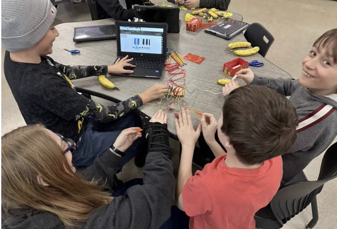 A group of students gather around a laptop, Makey Makeys, and various items to attach for exploration. 