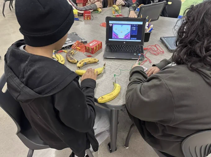 Two students work with bananas attached to Makey Makeys on their laptop. 