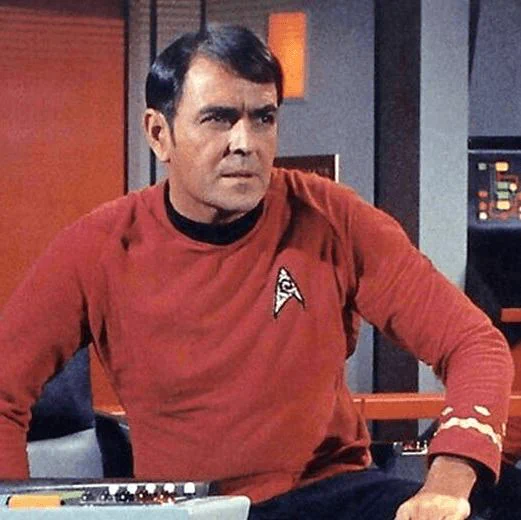 James Doohan as Scotty sits at a control console aboard the USS Enterprise, wearing a red Starfleet uniform with a black collar and a delta insignia on his chest. He leans slightly forward with one hand resting on the console, his dark hair neatly combed to the side. Behind him are colorful control panels and futuristic set walls characteristic of the original Star Trek series bridge.