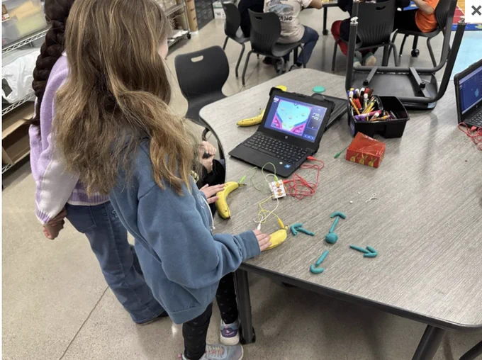 Students gather around a laptop with Makey Makeys, bananas, and Play-Doh, exploring various features of the technology. 