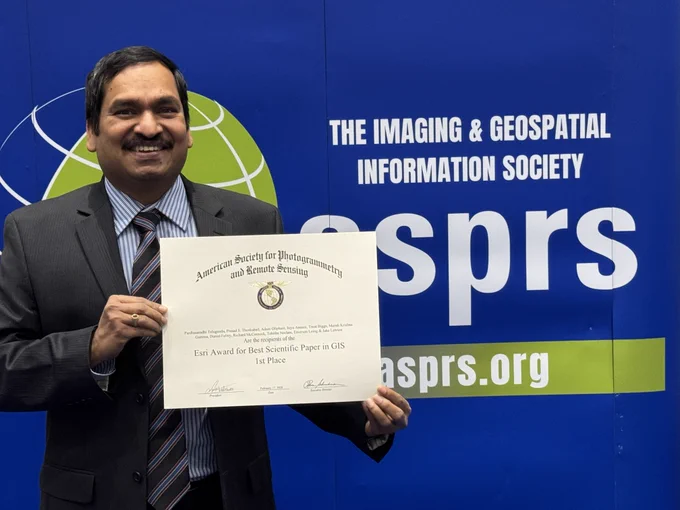 Dr. Teluguntla holds the award certificate he received for his team while standing in front of an American Society of Photogrammetry and Remote Sensing banner.