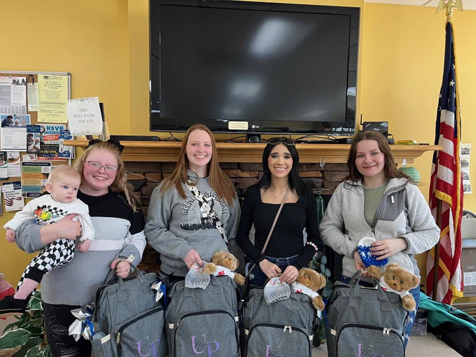 Four women pose with baby, backpacks, and teddy bears. The backpacks have \"UP\" on them. The woman on the left holds a baby in a white race car outfit. An American flag is on the right. Text reads: FREE WELL WATER TEST KITS.
