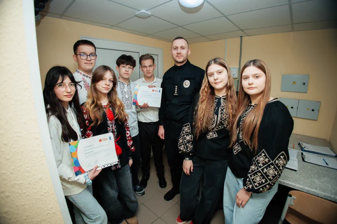 A group of secondary school students stand alongside a juvenile prevention officer inside a school office in Zaporizhzhia, holding certificates after completing mediation and non-violent communication training. The students wear traditional embroidered shirts and casual attire, while the officer stands in uniform.