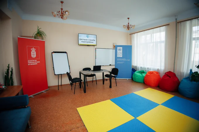 A specially equipped School Reconciliation Service classroom in Zaporizhzhia featuring a mediation space with chairs, a whiteboard and beanbags. Denmark and UNDP banners stand in the room, and a screen displays the words “School Reconciliation Service.” The safe, welcoming environment is designed to support dialogue, peer mediation and non-violent conflict resolution among students.