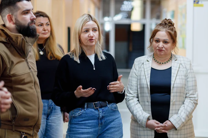 Four adults stand in a softly lit school corridor during the launch of the School Reconciliation Services initiative in Zaporizhzhia. A woman wearing a black sweater and blue jeans gestures with her hands as she talks. Her expression is focused and engaged. Standing beside her are partner representatives, listening attentively.