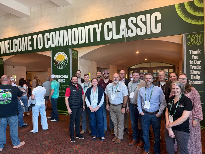 A group of approximately 15 individuals stands in an entrance hallway beneath a sign that reads "Welcome to Commodity Classic".