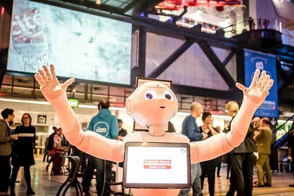 Un robot humanoïde accueille les visiteurs, bras levés, au cœur d'un espace d'exposition animé. autour de lui, des stands, des écrans géants et des passionnés échangent sur les sciences et les technologies.