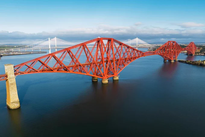 The red  Forth Bridge with Forth Road Bridge and Queensferry Crossing behind, over the dark blue river Forth and blue skies above.  
