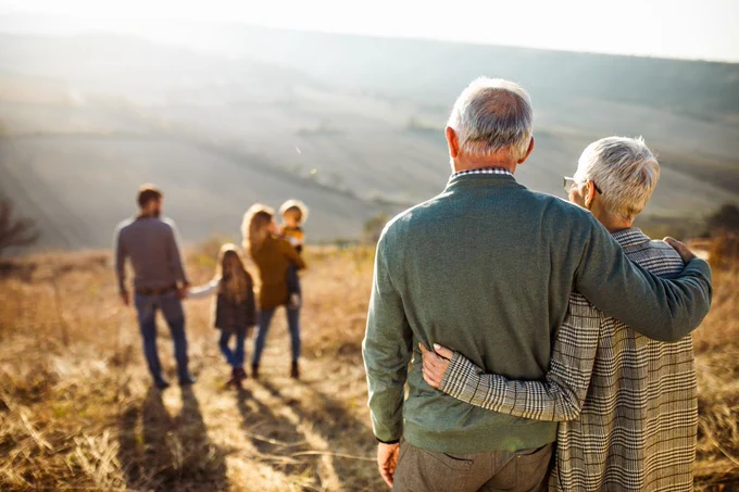 Two older adults, one with a blanket over their shoulders, watch two younger adults walk hand in hand through a rolling countryside landscape during sunset.