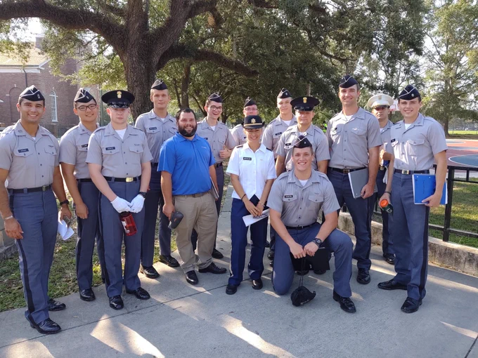 A group of cadet students from The Citadel in uniform and a person in a blue shirt are gathered outside under a large tree. Ten individuals are wearing military-style uniforms and caps, and three are in civilian attire. Dimitra Michalaka is wearing a white uniform with blue pants. They are standing on a sidewalk with buildings and trees in the background. Some are holding books and a water bottle. The mood is relaxed and friendly.