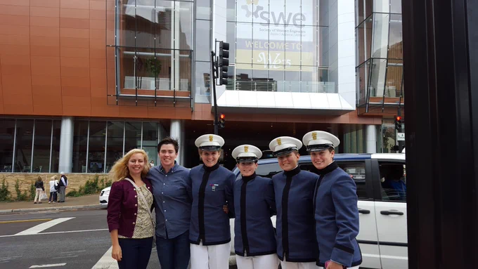 Dimitra Michalaka at the far left with female cadets from The Citadel stand on a city street in front of a building with a sign that reads "SWE Welcome to St. Louis." Five individuals wear uniforms and four of those with hats, while Dimitra is dressed in civilian attire. They are all smiling.