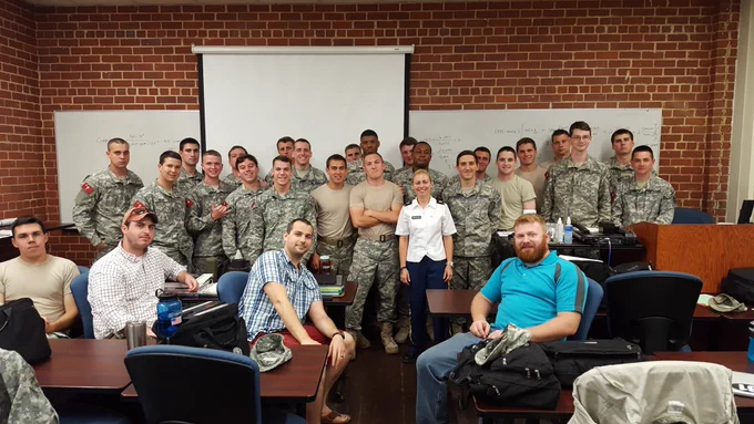 A group of cadet students from The Citadel in military uniforms and civilian clothing are gathered in a classroom. Dimitra is in her white shirt and blue pant uniform and stands in the middle of the group. They are posing for a photo in front of a whiteboard and a projector screen. Some are seated at desks, while others are standing. The room is decorated with brick walls.