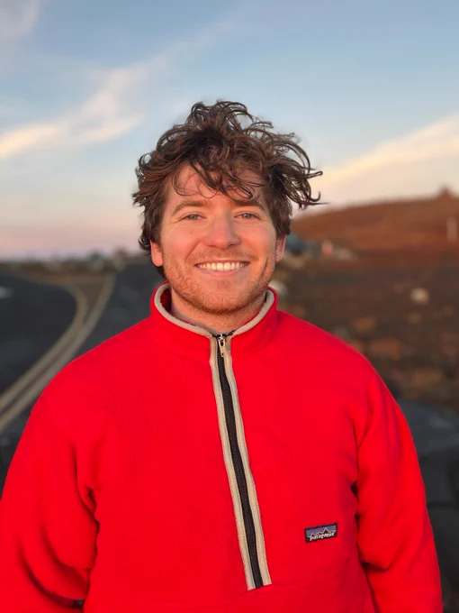 Photograph of a man wearing a red fleece coat pictured in front of a road and field at sunset. 𝘗𝘩𝘰𝘵𝘰 𝘣𝘺 𝘔𝘢𝘳𝘪𝘯𝘢 𝘚𝘢𝘯𝘵𝘰𝘴.