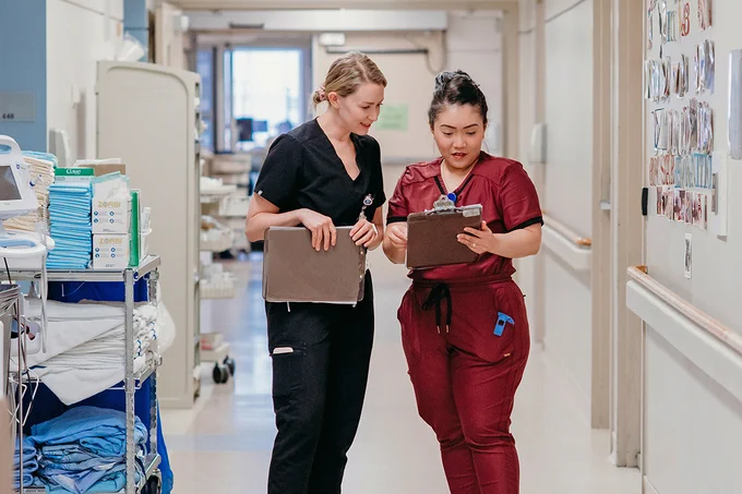 Two healthcare professionals walking down a hospital corridor, both holding clipboards. One is dressed in black scrubs, the other in maroon. Shelves with medical supplies and linens are seen on the left, and there's a display of images on the right wall.