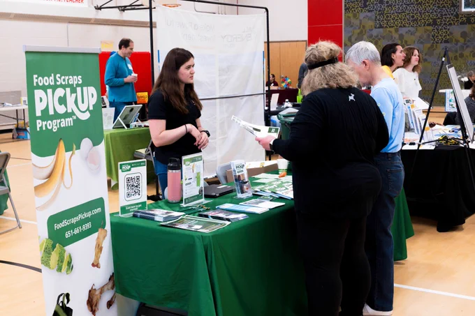 person stands behind a table with green table cloth and printed materials with two people on opposite side looking at materials at an expo