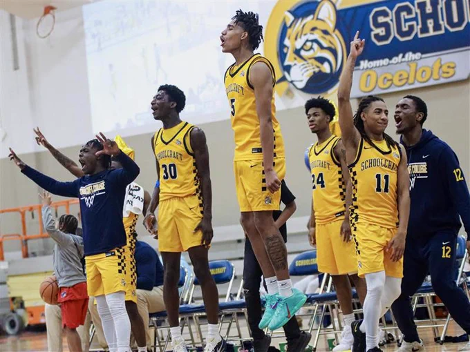 A group of basketball players in yellow uniforms, displaying energetic reactions on the sidelines. They are standing or jumping near a bench, with some raising their arms. The gym features a large wall display showing an ocelot logo and the words "Schoolcraft Ocelots."
