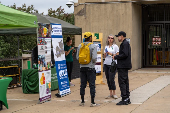 A table set up with information about the Central Avenue Blue/Silver Line plan, and people discussing around it. 