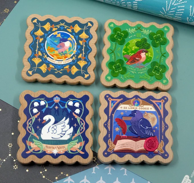 A photograph of a set of 4 wooden magnets, shaped like square wafers with embossed bird designs. The designs featured are: ginkgo leaves and an Iberian magpie, a sparrow and clovers, a fairytale swan and lily-of-the-valley flowers, and a jackdaw in a library modeled after an ex libris.
