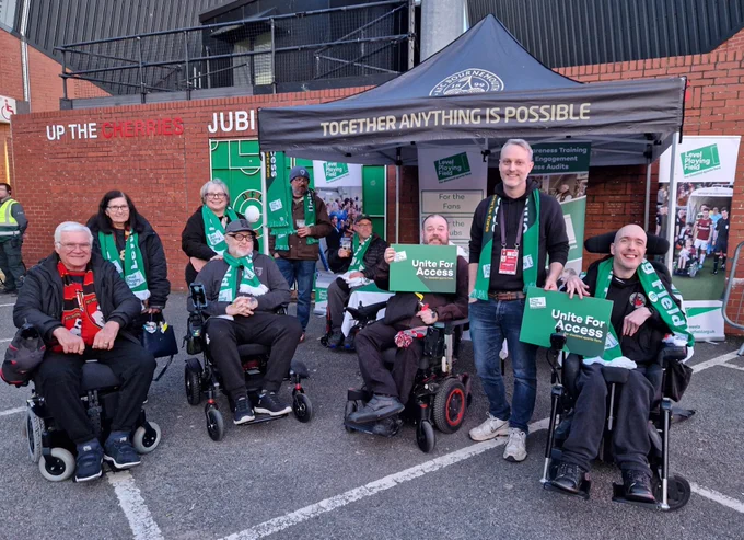 Level Playing Field’s Daniel Townley is joined by AFC Bournemouth Disabled Supporters’ Association members, smiling in front of Level Playing Field banners and a black marquee with the text “Together Anything Is Possible.” Many are wearing green Level Playing Field scarves and two people at the front are holding Unite For Access boards.