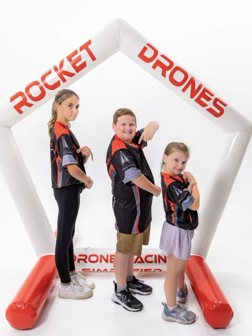 Three individuals are posing confidently in front of a white and red inflatable arch with the words "Rocket Drones" and "Drone Racing." They are wearing matching black and red jerseys and are standing on a white floor.