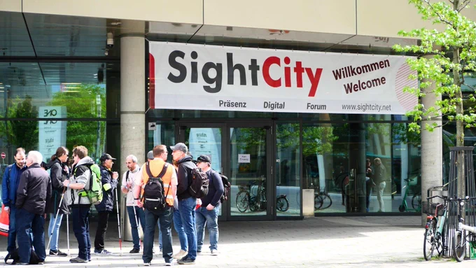 People, some with canes, outside a building with a banner reading Sight City with Wilkommen, Welcome and Präsens Digital Forum www.sightcity.net