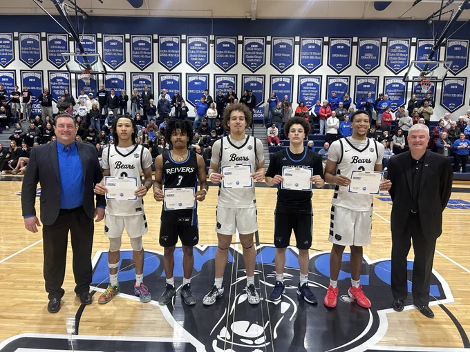 Seven people stand on a basketball court for an awards photo: two presenters in suits at each end and five players in the middle holding certificates. Three players wear white “Bears” uniforms, and two wear dark “Reivers” uniforms. A packed crowd and a wall of blue championship banners fill the background.