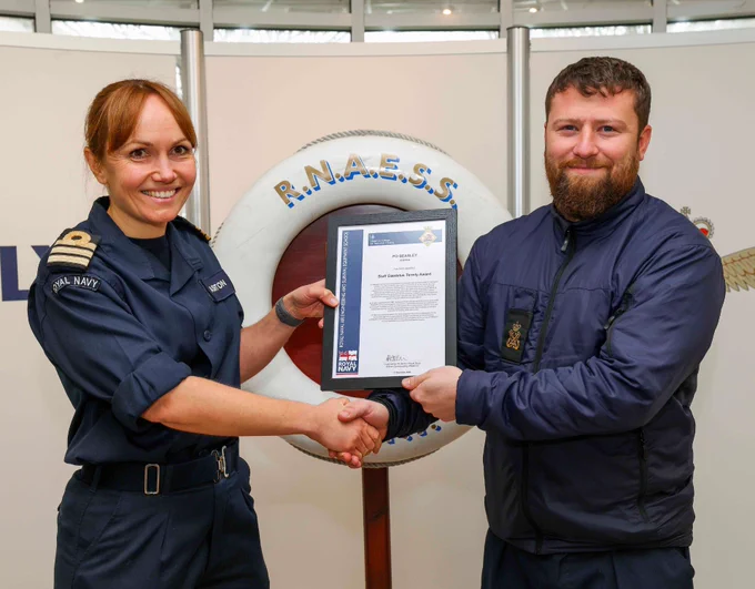 The image shows two people shaking hands. One is wearing a navy uniform, and the other is holding a certificate. There is a Royal Navy badge on the uniform, and a life preserver with "R.N.A.E.S.S." behind them.