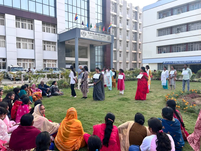 A group of women sit in a circle outdoors on a lawn, watching others stand in lines holding placards in front of a large institutional building.