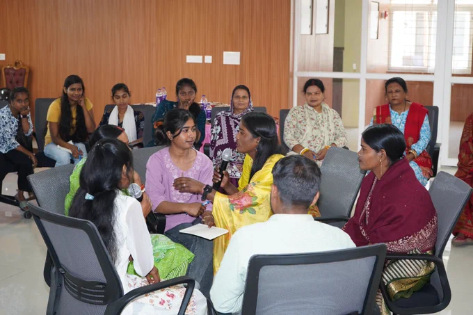 Women on chairs, sit in a close circle indoors during the training, with one woman speaking into a microphone while others listen attentively.