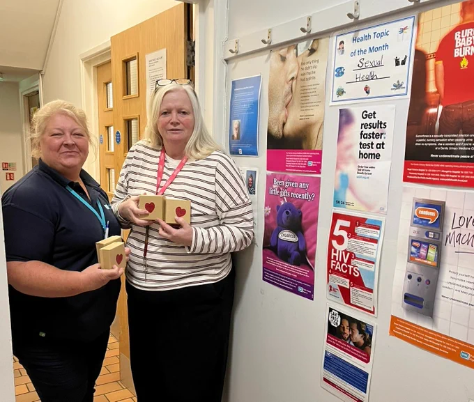 Homeless Health Nurse Sharon Graham with a hostel staff member. They are standing in front of an information board displaying info about sexual health and are holding small boxes with a heart on them.