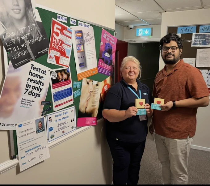 Homeless Health Nurse Sharon Graham with a hostel staff member. They are standing in front of an information board displaying info about sexual health and are holding small boxes with a heart on them.
