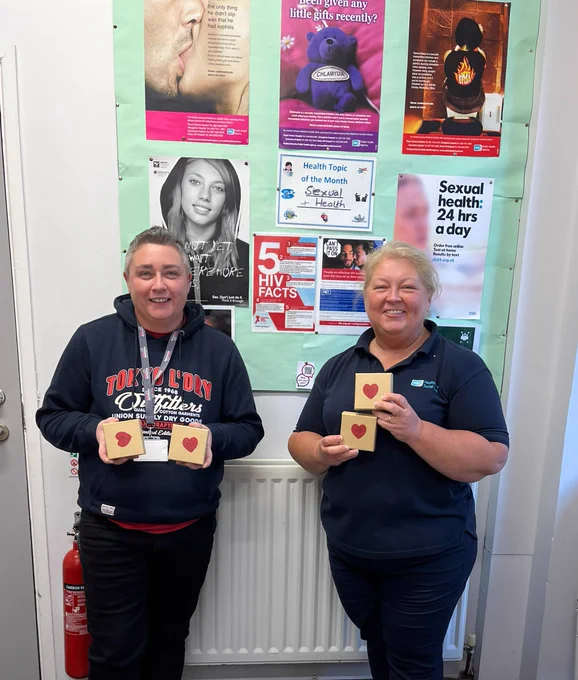 Homeless Health Nurse Sharon Graham with a hostel staff member. They are standing in front of an information board displaying info about sexual health and are holding small boxes with a heart on them.