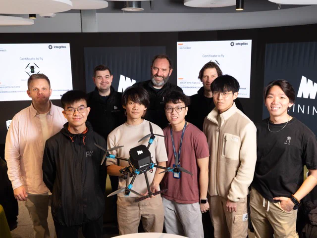 Eight people pose indoors in casual attire. Two hold a small drone with blue accents. Behind them, screens display a logo and the words "Certificate to fly." 