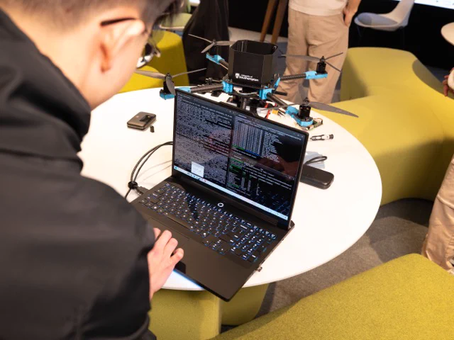 A person types on a black laptop displaying code, with a drone resting on a round table in front of them. The setting is informal, featuring green cushioned seating in the background.