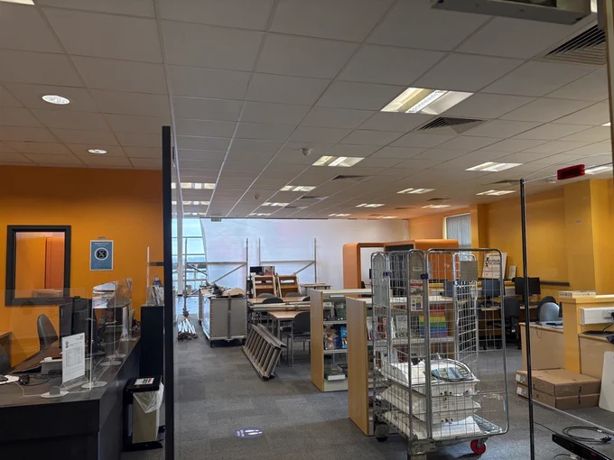 Interior view of the Walsall Manor Hospital library on Route 302, currently closed for essential ceiling repair work. The space shows orange walls, ceiling tiles, scattered chairs and tables pushed aside, mobile metal book trolleys filled with books, construction scaffolding and white protective sheeting erected in the background, computer workstations and various library materials in disarray during the refurbishment.
