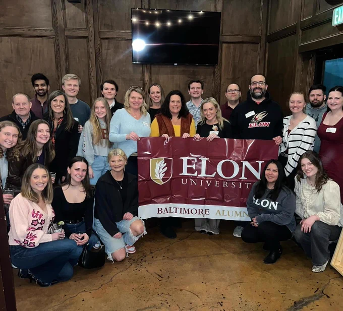 A group of people standing indoors, posing with a banner that reads "Elon University Baltimore Alumni." Some individuals are holding beverages. The setting is a wood-paneled room with a wall-mounted TV.