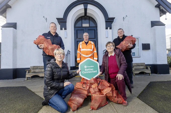 Group of people standing and kneeling outside a white building, holding and surrounded by multiple red net bags filled with firewood. A sign reading \u2018Translink Better.Connected.\u2019 is displayed in front.