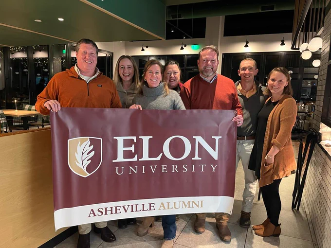 A group of people standing indoors, holding a large banner that reads "Elon University Asheville Alumni." They are smiling and posing for the photo in a modern interior setting.