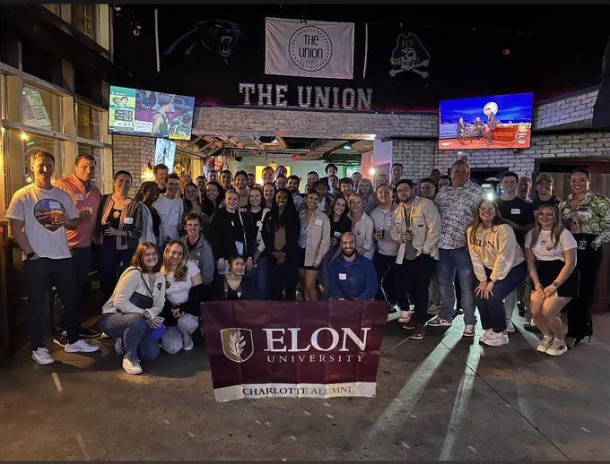 A group of people are gathered inside a venue named "The Union." They are posing for a group photo, holding a banner that says "Elon University Charlotte Alumni." The room has several sports-related items on the walls and screens showing events.