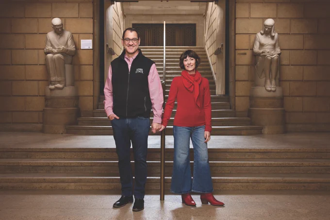 President David Cook and his wife Katie stand holding hands at the base of a grand stone staircase inside Parks Library. Flanking the stairs on either side are larger than life limestone sculptures created in 1944 by Christian Petersen, known as The Library Boy and The Library Girl, each seated and reading atop a pedestal. Cook wears glasses, a pink button down shirt and a black Iowa State vest, and Katie wears a red sweater, scarf, jeans and red boots.