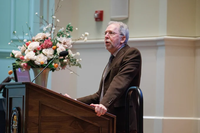 Eric Green, the keynote speaker, stands at a wooden lectern wearing a brown suit and glasses, speaking next to a large floral arrangement.