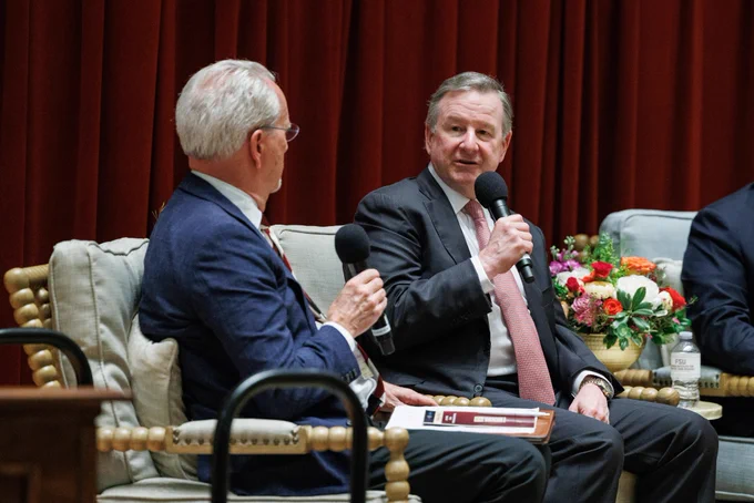 Richard McCullough, wearing a dark suit and light red tie, speaks into a microphone while seated between other panel members on stage.