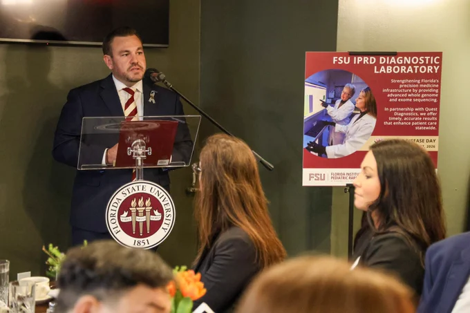 Rep. Adam Anderson speaks at a Florida State University lectern, discussing the FSU IPRD Diagnostic Laboratory beside a promotional poster for the facility.