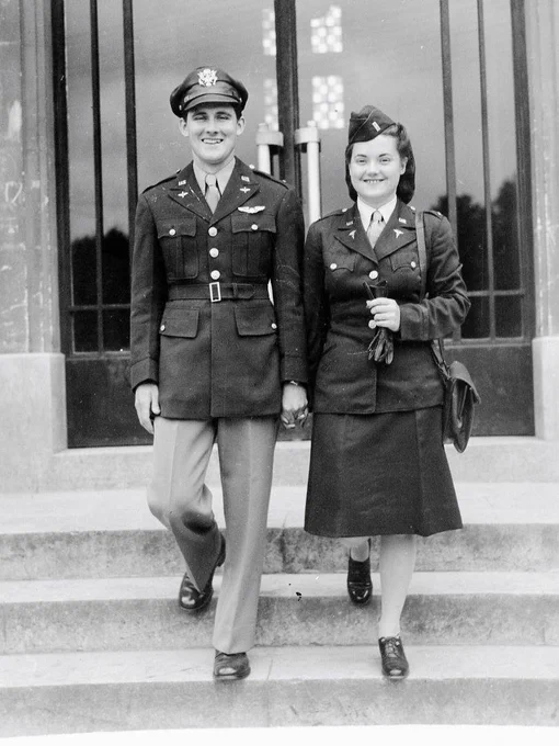 Two people in military uniforms descending steps, smiling. The uniforms display insignia and badges. They are in front of a building with glass doors.