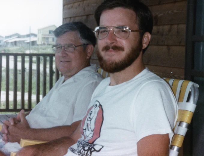 Two people sit on a porch in front of a brown house. They are both in lawn chairs, wearing glasses, and smiling.