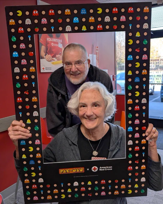 Two individuals are smiling and posing inside a large frame decorated with Pac-Man graphics. The frame features branding for both Pac-Man and the American Red Cross. In the background, a poster with the word "volunteers" is partially visible on a red wall.