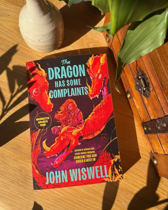 Advanced paperback copy of The Dragon Has Some Complaints by John Wiswell on a table next to a chest, vase, and plant.