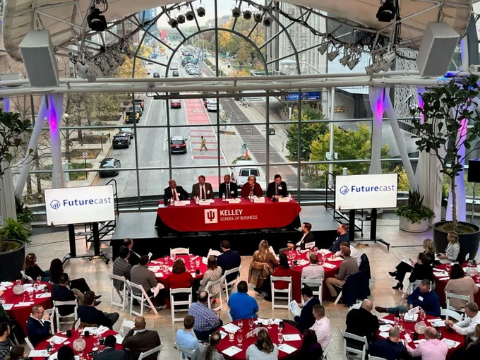 A group of individuals seated at a head table on a stage with a red tablecloth marked "Kelley School of Business." Two screens display the "Futurecast" logo. The audience sits at round tables with red tablecloths in a glass-walled venue overlooking a street.